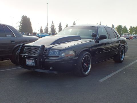 2003 Ford Crown Victoria - black full-size sedan modified with a chrome push-bar grille guard, halo projector headlights and a hood scoop, parked at a car meet.