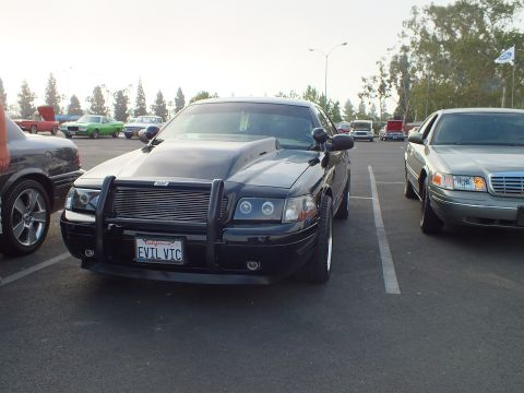 2003 Ford Crown Victoria - Front view of a black full-size sedan fitted with a chrome push bar and a license plate reading EVIL VIC, parked among other cars.