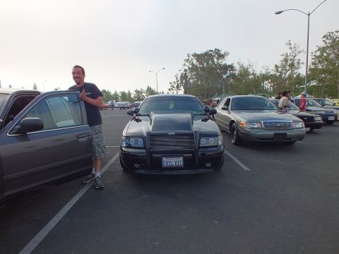 2003 Ford Crown Victoria - smiling man stands by an open car door next to a black sedan with a hood scoop and EVIL VIC plate at an outdoor car meet.