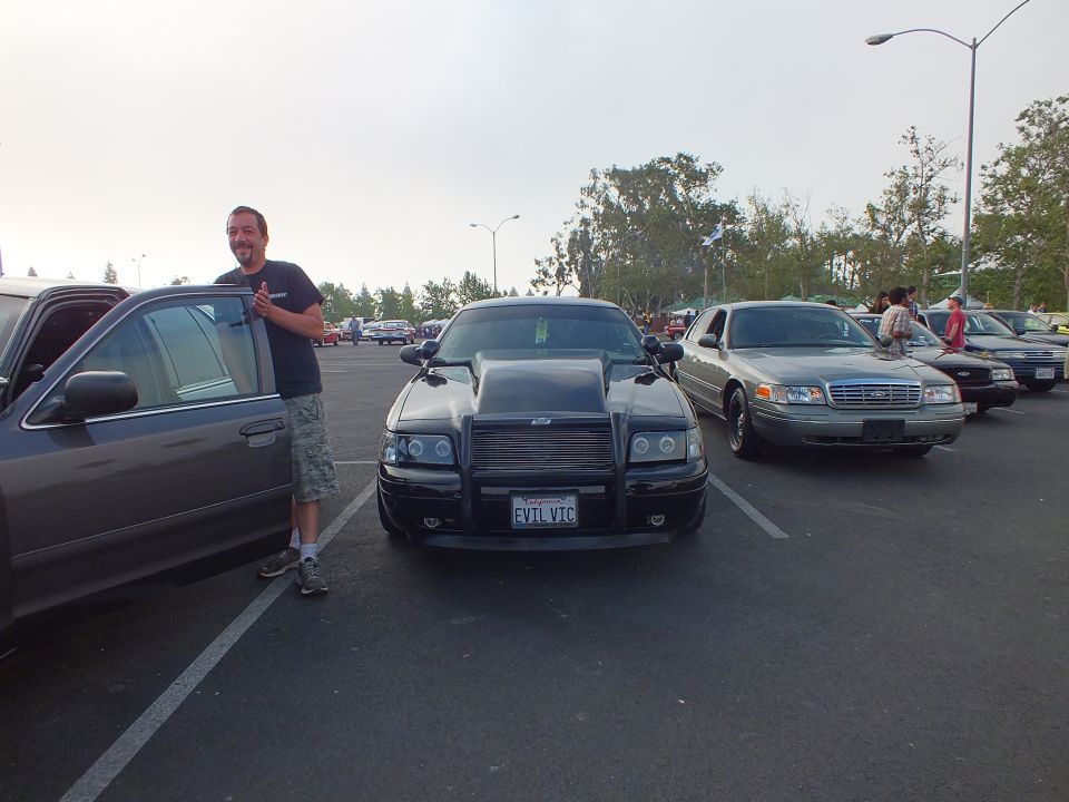 Man standing beside a black sedan with hood scoop and EVIL VIC plate in parking lot