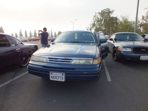 2003 Ford Crown Victoria - blue Ford sedan seen from the front with the oval Ford badge and a California license plate reading NARCPTR, parked at a meet.