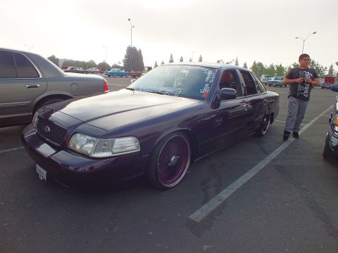 2003 Ford Crown Victoria - lowered dark purple Ford sedan on pink wire-spoke wheels sits beside a standing man at an outdoor car meet.