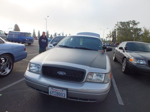 2003 Ford Crown Victoria - silver Ford full-size sedan with a blacked-out grille and California plate 6YIB803, with a man standing behind it at a meet.