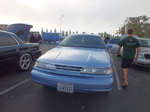 2003 Ford Crown Victoria - light blue Ford full-size sedan viewed head-on with a California plate reading 1SWTVIC, surrounded by other cars and people.