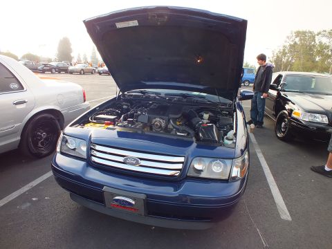 2003 Ford Crown Victoria - dark blue Ford full-size sedan with its hood raised, revealing the engine bay, parked between other cars at a meet.