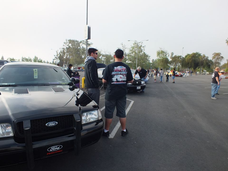 Two men stand by a black push-bar sedan at a parking-lot car meet