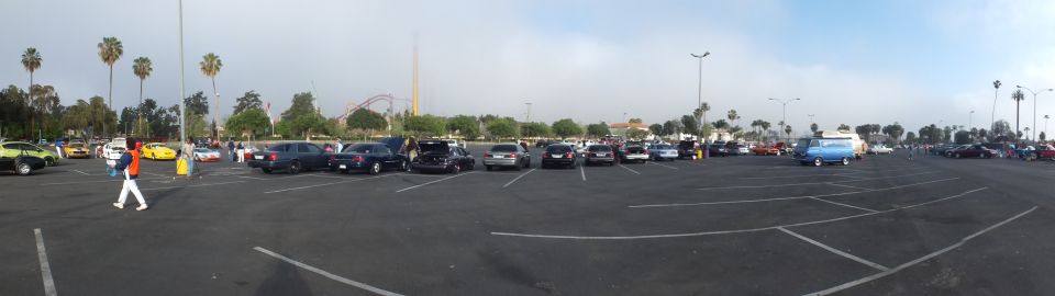 Panorama of parked cars at a meet with people and a roller coaster in distance