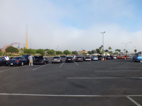 2003 Ford Crown Victoria - row of parked cars sits in a wide lot with a tall smokestack and roller coaster visible in the distance under cloudy sky.