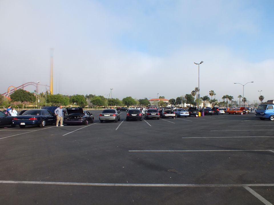 Row of cars in parking lot with smokestack and roller coaster in distance