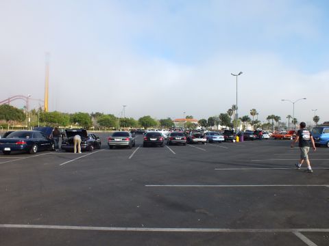 2003 Ford Crown Victoria - parking-lot car meet with several parked cars, a man walking across the asphalt, and a roller coaster in the background.