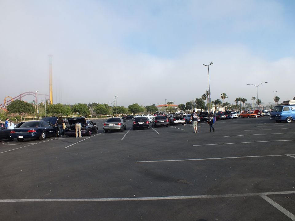 Wide parking lot with parked cars and people near a roller coaster horizon