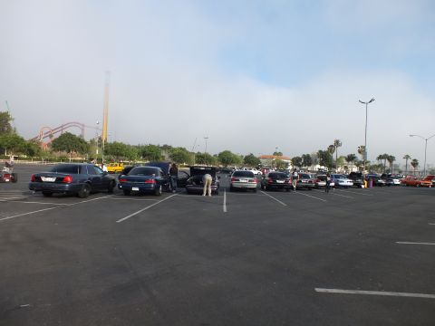 2003 Ford Crown Victoria - Cars are parked across a large lot as people load open trunks, with a roller coaster visible in the distance under a hazy sky.