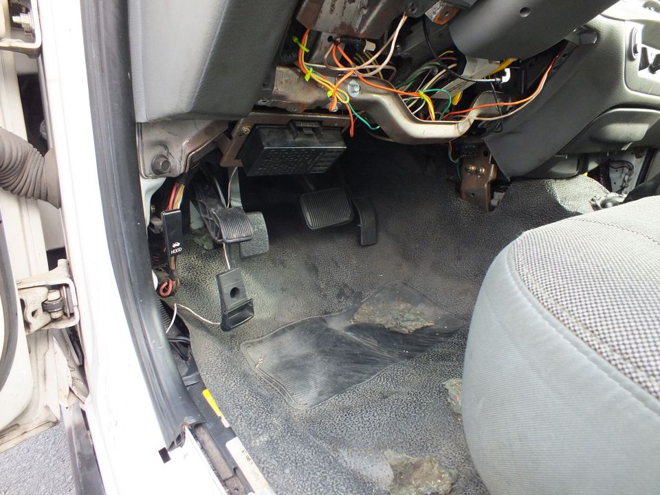 Driver footwell of a car with exposed wiring and pedals on gray carpet