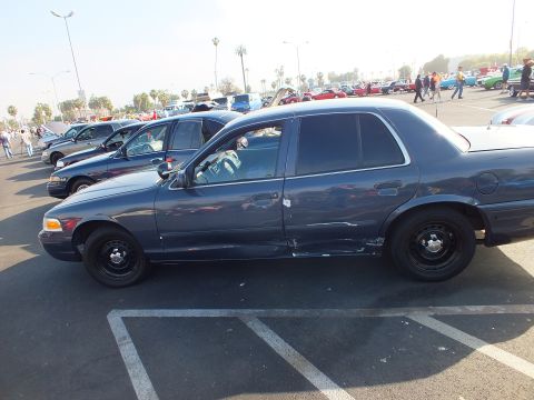 2003 Ford Crown Victoria - Side profile of a dark blue Ford sedan on black steel wheels parked among many cars at an outdoor car show.
