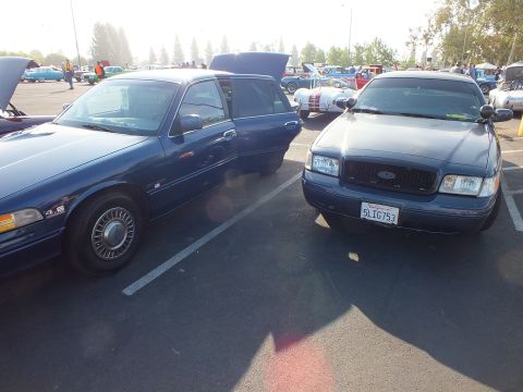2003 Ford Crown Victoria - Two blue Ford sedans parked facing one another at an outdoor show; the right car shows California plate 5LIG753 and a Ford grille emblem.