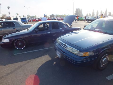 2003 Ford Crown Victoria - lowered dark blue Ford sedan on chrome wire wheels parked beside a navy Ford with its hood raised at an outdoor car show.
