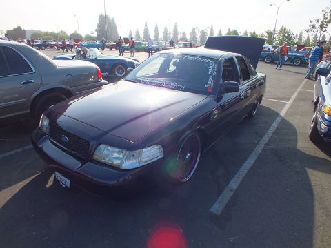 2003 Ford Crown Victoria - dark purple lowered Ford sedan with hood and trunk open, custom wheels and windshield decals, parked at an outdoor car show.
