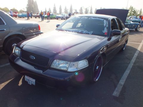 2003 Ford Crown Victoria - Front three-quarter of a dark purple lowered Ford sedan with custom wheels, hood up and 'Lv Customs' and 'Venom' windshield lettering.
