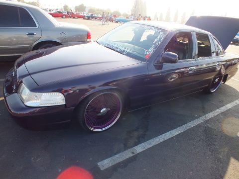 2003 Ford Crown Victoria - Side profile of a dark purple lowered Ford sedan riding on purple-laced wire wheels, hood and trunk open at an outdoor show.