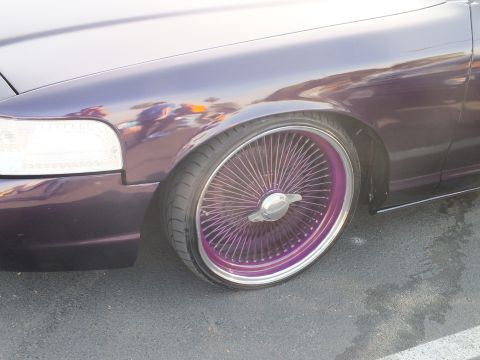 2003 Ford Crown Victoria - Close-up of a glossy purple car's front fender and tire wrapped around a purple-spoked chrome wire wheel.