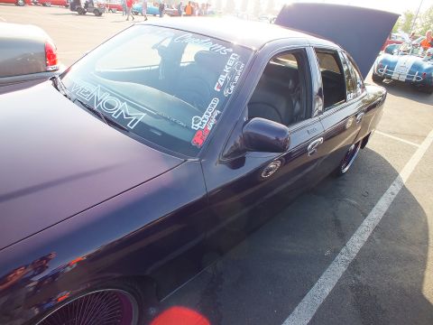 2003 Ford Crown Victoria - Rear three-quarter of a glossy dark purple sedan with windshield decals reading VENOM and Falken, trunk open at a car show.