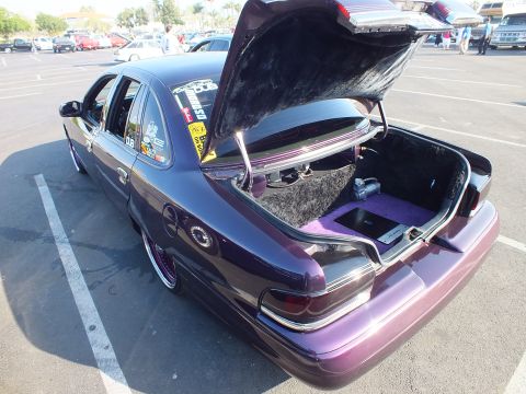 2003 Ford Crown Victoria - Rear three-quarter of a lowered purple sedan with its trunk open, revealing a custom purple-carpeted setup at an outdoor show.
