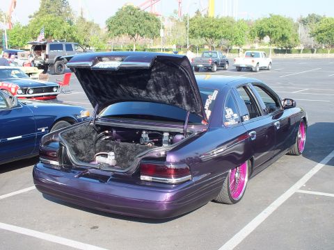 2003 Ford Crown Victoria - purple metallic full-size sedan sits low on pink-spoke wire wheels with its carpeted trunk propped open in a sunny show lot.