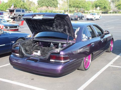 2003 Ford Crown Victoria - rear of a purple metallic sedan with its trunk lid open, riding low on pink wire-spoke wheels among other show cars.