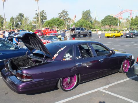 2003 Ford Crown Victoria - purple metallic sedan sits slammed to the ground on pink wire wheels, trunk open, with a roller coaster visible behind the show field.