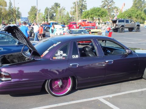 2003 Ford Crown Victoria - lowered purple metallic four-door sedan sits on pink wire-spoke wheels, its side windows covered in decals, at an outdoor meet.