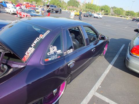 2003 Ford Crown Victoria - rear quarter glass of a purple sedan is plastered with decals such as Falken and DUB, with pink wire wheels visible below.