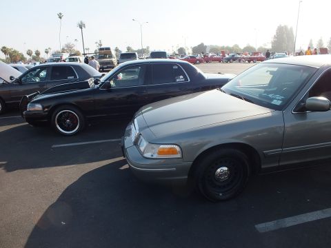 2003 Ford Crown Victoria - gray full-size sedan and a black sedan sit nose-out in a show lot, palm trees and other cars in the background.