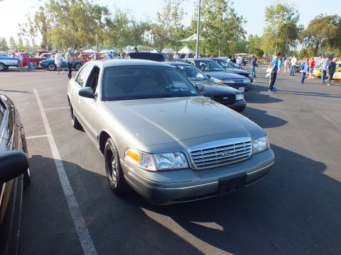 2003 Ford Crown Victoria - gold-gray Ford Crown Victoria sedan with chrome grille and Ford emblem sits in a show field with crowds and tents behind.