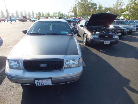 2003 Ford Crown Victoria - silver Ford sedan faces the camera with its Ford-oval grille and California plate 6YIB803, a dark Ford with open hood beside it.