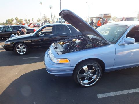 2003 Ford Crown Victoria - light blue Ford full-size sedan sits with hood raised on large chrome wheels, parked next to a black sedan at the car show.