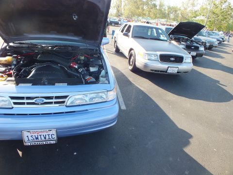 2003 Ford Crown Victoria - light blue Ford sits with hood up showing a red intake and plate 1SWTVIC, a silver Ford Crown Victoria parked alongside.