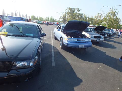 2003 Ford Crown Victoria - light blue Ford sedan with its hood raised sits between a black sedan and a silver Crown Victoria in a busy show parking lot.