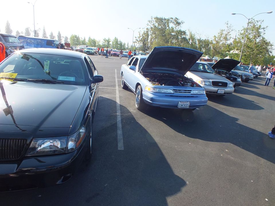 Light blue Ford sedan with open hood beside a black sedan and silver Crown Victoria at a show
