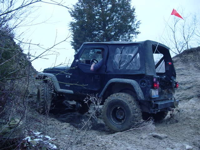 Black Jeep Wrangler with soft top driving on a muddy, rocky trail with trees in the background