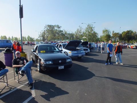 2003 Ford Crown Victoria - black Mercury Grand Marquis sits in a sunny parking-lot car show as a man relaxes in a folding chair nearby.