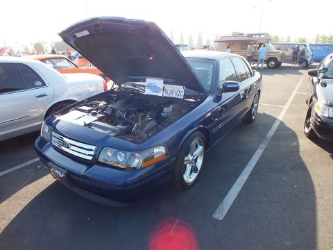 2003 Ford Crown Victoria - dark-blue Ford sedan displays its engine with the hood up and a hand-lettered BLUE VIC sign at a car show.