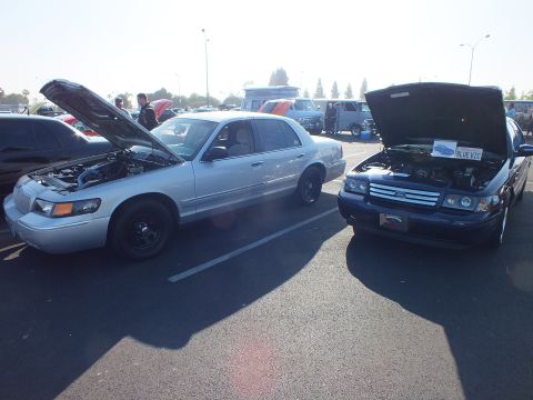 2003 Ford Crown Victoria - silver Mercury Grand Marquis and a blue Ford sedan sit nose to nose with hoods raised at a car show.