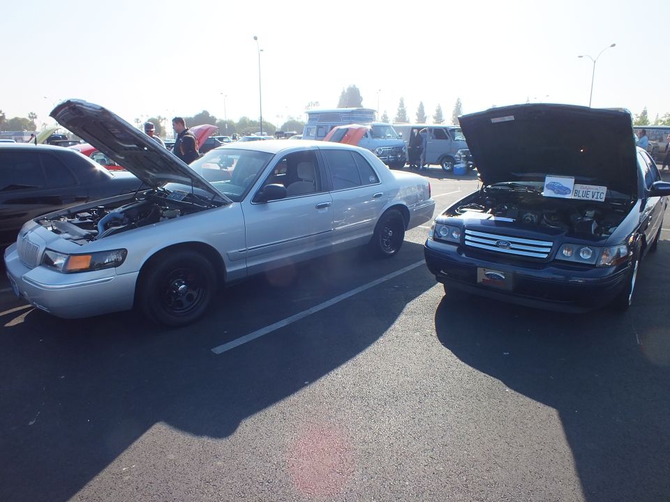 Silver Mercury and blue Ford sedan parked nose-to-nose with hoods raised at a car show