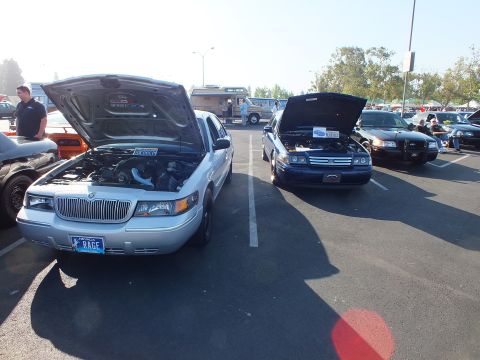 2003 Ford Crown Victoria - silver Mercury Grand Marquis with hood up and a RAGE license plate sits beside a blue Ford at a car show.