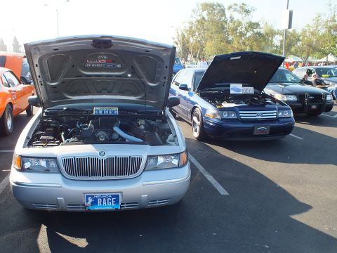 2003 Ford Crown Victoria - silver Mercury with a Nevada RAGE plate and a blue Ford sedan display their engines, hoods raised, at a car show.