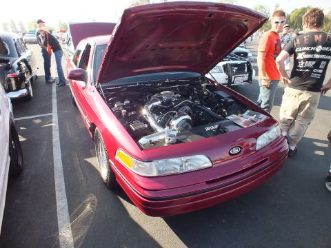 2003 Ford Crown Victoria - red Ford sedan shows a supercharged engine with its hood up while spectators gather around at a car show.