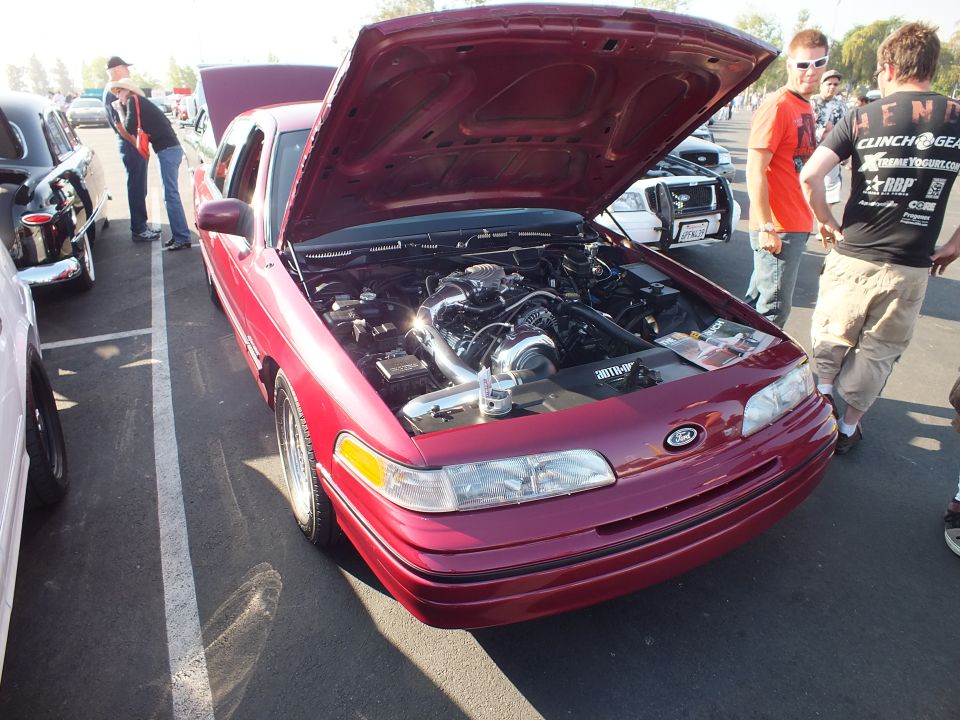 Red Ford sedan with open hood and supercharged engine as spectators look on at a car show