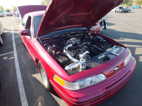 2003 Ford Crown Victoria - red Mercury sedan displays a supercharged engine with the hood up in a sunny parking-lot car show.