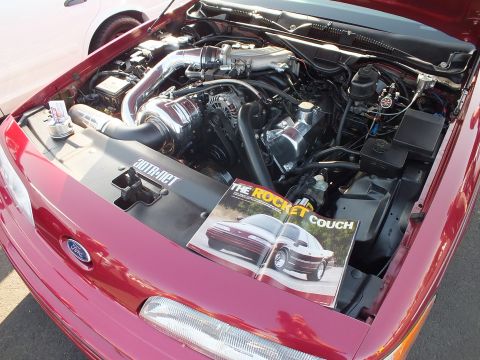 2003 Ford Crown Victoria - supercharged engine bay of a red Ford with a Vortech-style blower, photographed beside an open magazine spread titled 'The Rocket Couch'.
