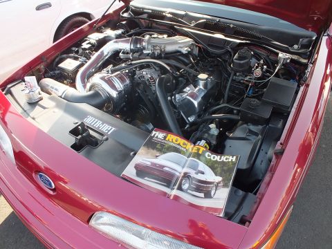 2003 Ford Crown Victoria - Close view of a red Ford's supercharged engine bay with a magazine propped open showing a feature titled 'The Rocket Couch'.